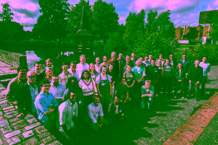 Group photo of the workshop participants outdoors on the University of Twente campus, standing beside a pond with a small brick tower.
